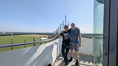 Auf einem Aussichtsturm stehen zwei Jungs, die für das Foto die Arme umeinander gelegt haben. Im Hintergrund sind Windräder erkennbar, der Himmel ist wolkenlos.