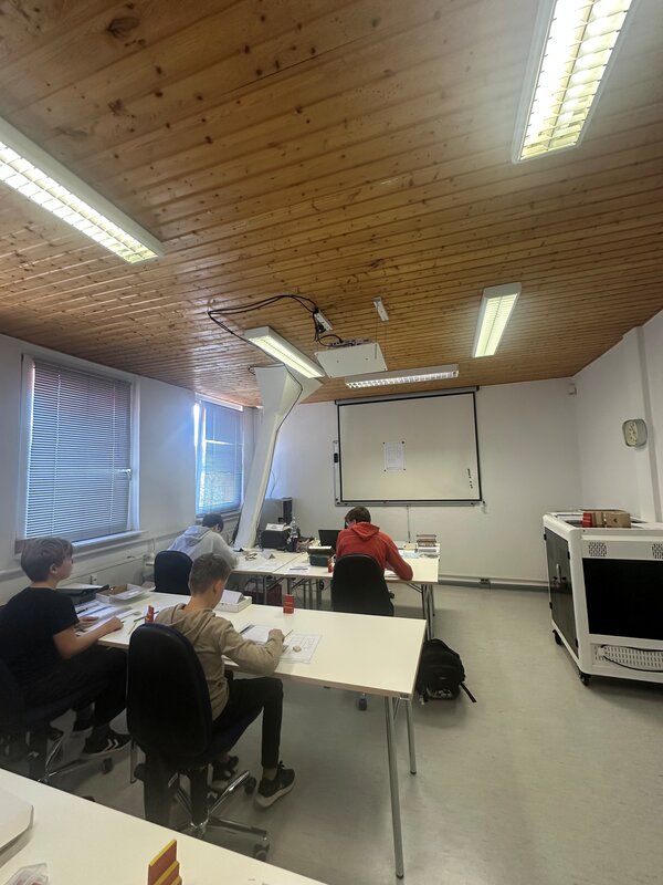 Mehrere Schüler arbeiten in einem Unterrichtsraum konzentriert an Aufgabenblättern und kleinen Werkstücken. Im Raum befinden sich eine Projektionsfläche, ein Beamer und technische Arbeitsmaterialien.