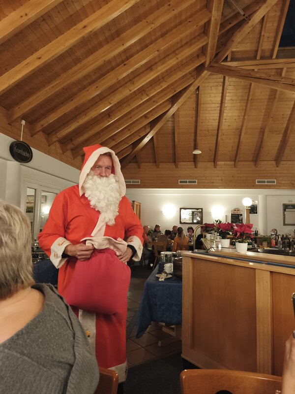 Nikolaus steht mit rotem Geschenksack vor dem Tresen einer Gastwirtschaft unter holzvertäfelter Decke; Gäste sitzen im Hintergrund.