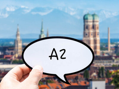 Eine Hand hält eine Sprechblase mit der Aufschrift 'A2' in den Vordergrund. Im Hintergrund ist die Skyline von München mit markanten Gebäuden wie dem Frauenkirche zu sehen. Die Stadtansicht zeigt eine Mischung aus historischen und modernen Gebäuden unter einem leicht bewölkten Himmel.