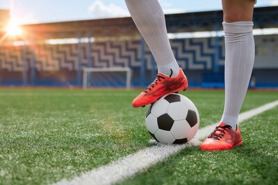 Eine Frau in Fußballbekleidung (das heißt: Fußballschuhe und Stulpen) steht mit einem Bein auf einem schwarz-weißen Lederfußball, der auf der Seitenlinie eines Fußballplatzes in einem Stadion liegt. Mann sieht die Frau nur bis zu den Knien. Im Hintergrund steht die Abendsonne tief hinter der Tribüne.