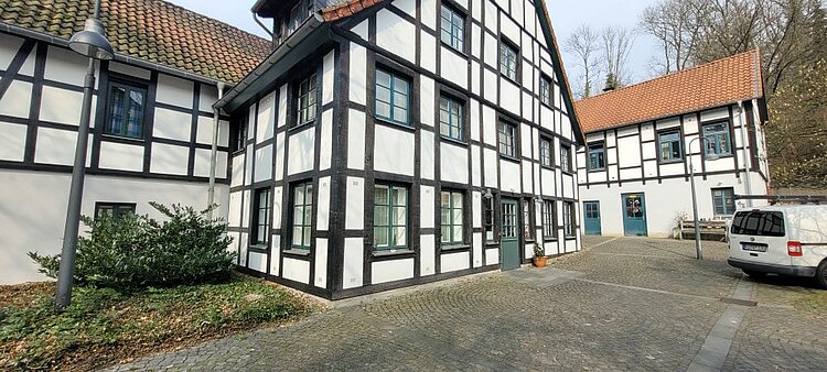Ein historisches Fachwerkhaus mit weißem Fächern, braunen Balken, blauen Fensterrahmen und roten Dachziegeln. Im Hintergrund ist ein Nebengebäude zu sehen. Auf dem Hof steht ein Lieferwagen.