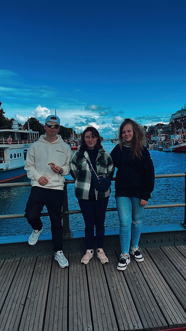 Zwei Schülerinnen und ein Schüler stehen auf einer Brücke über einen Fluss und posieren dort. Im Hintergrund sind rechts und links Häuser.