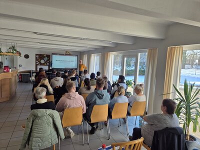 In einem gefliesten Raum sitzen in mehreren Stuhlreihen etwa 20 Personen, die dem Vortragenden lauschen, der ganz vorne neben einem großen Bildschirm steht, auf dem der Text "Brandschutzschulung 2025" geschrieben ist. Links im Raum befindet sich eine Theke mit Kaffeekannen darauf, rechts führt der Blick durch die bodentiefen Fenster in einen verschneiten Garten.