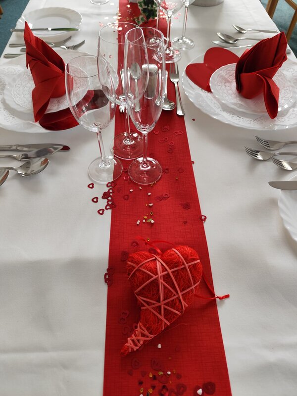 Eine stilvolle Tischdekoration mit einem roten Läufer, roten Servietten und transparenten Weingläsern. In der Mitte liegt ein dekoratives, geflochtenes rotes Herz.