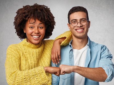 Zwei lächelnde Personen, eine Frau mit lockigen Haaren und ein Mann mit Brille, stehen nebeneinander und machen eine Faust-an-Faust-Geste. Die Frau trägt einen gelben Pullover, der Mann ein hellblaues Hemd. Der Hintergrund ist neutral und grau.