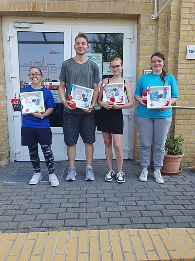 Geschafft! Vor der Tür der Euro-Schulen stehen drei zufrieden grinsende Schülerinnen und ein Schüler. Sieh halten individuell gestaltete, weiße Bilderrahmen in der Hand, sowie verschiedene kleine Geschenke.