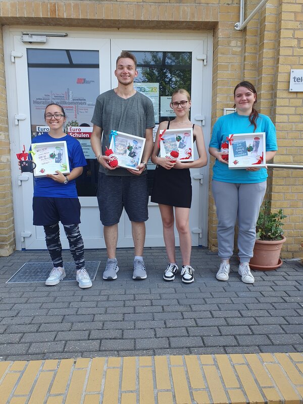 Vor der Tür der Euro-Schulen stehen drei zufrieden grinsende Schülerinnen und ein Schüler. Sieh halten individuell gestaltete, weiße Bilderrahmen in der Hand, sowie verschiedene kleine Geschenke.