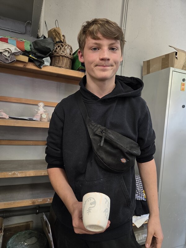 Ein junger Mann in schwarzem Kapuzenpulli präsentiert eine selbst bemalte Tasse. Auf einem Regal im Hintergrund sind weitere Bastelarbeiten zu sehen.