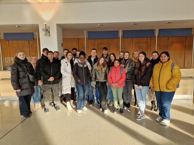 Ein Gruppenbild in dem Foyer der Messehalle in Leipzig. Die Personen tragen dicke Jacken und einige tragen Rucksäcke. Sie lächeln in die Kamera.