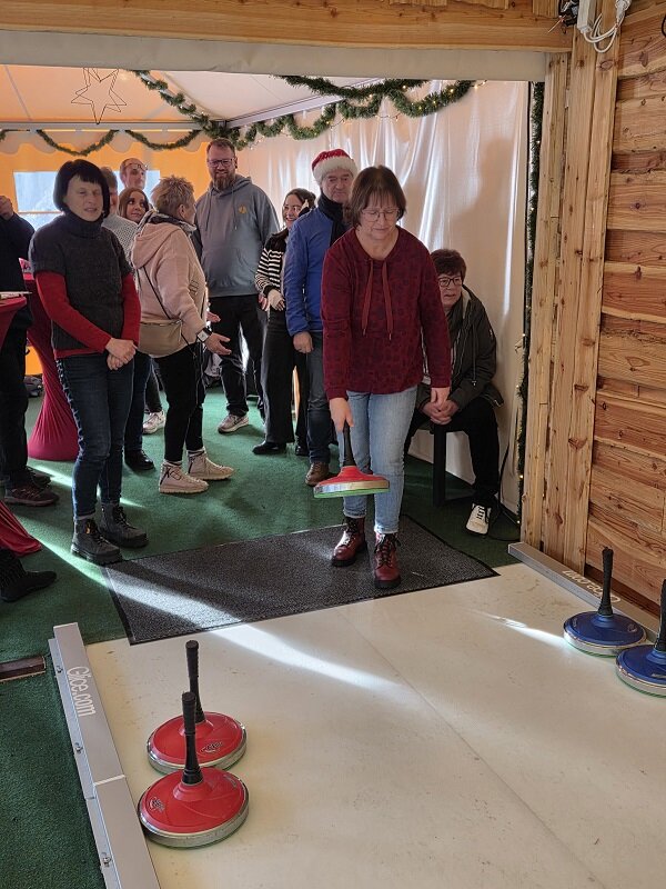 Eine Frau hält einen roten Eisstock, während mehrere Personen im Hintergrund zuschauen und warten.