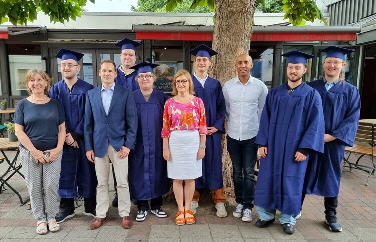 Gruppenbild der Absolvent*innen in einem Biergarten unter großen Bäumen. Ein Teil der Personen ist in dunkelblaue Talare gehüllt und trägt Absolventenhüte.