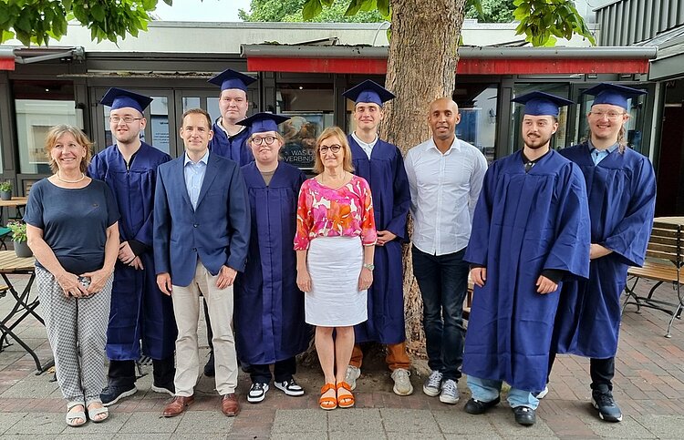 Gruppenbild der Absolvent*innen in einem Biergarten unter großen Bäumen. Ein Teil der Personen ist in dunkelblaue Talare gehüllt und trägt Absolventenhüte.