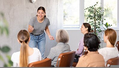 Pilotprojekt erfolgreich gestartet Eine junge Frau in grauem T-Shirt und Jeans spricht lebhaft vor einer Gruppe von Frauen, die auf Stühlen sitzen und ihr aufmerksam zuhören. Die Szene spielt in einem hellen Raum mit großen Fenstern und Pflanzen.