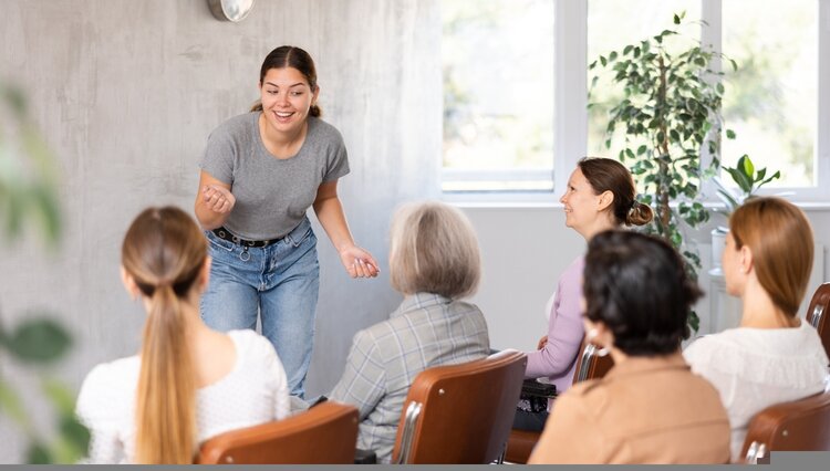Eine junge Frau in grauem T-Shirt und Jeans spricht lebhaft vor einer Gruppe von Frauen, die auf Stühlen sitzen und ihr aufmerksam zuhören. Die Szene spielt in einem hellen Raum mit großen Fenstern und Pflanzen.