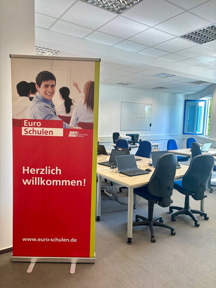 Blick in einen Unterrichtsraum, in dem gepolsterte Schreibtischstühle und Tische stehen und ein großes Euro-Schulen-Roll up an der Seite steht.