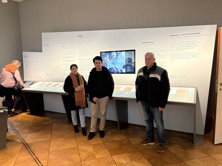 Drei Personen stehen in einem Ausstellungsraum vor einer langen Wand mit Texttafeln, Dokumentenvitrinen und einem Bildschirm mit Europakarte, während im Hintergrund weitere Besucher die Informationen der historischen Ausstellung lesen.