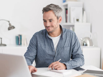 Ein Mann mit grauen Haaren sitzt an einem Schreibtisch und arbeitet an einem Laptop. Er trägt ein blaues Hemd und lächelt leicht. Auf dem Schreibtisch liegen ein Notizbuch und ein Smartphone. Im Hintergrund sind Bücherregale und eine Lampe zu sehen.