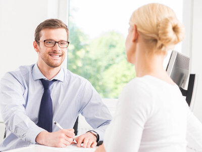 Ein Mann mit Brille und einem blauen Hemd sitzt an einem Schreibtisch und hält einen Stift in der Hand. Er lächelt freundlich und scheint ein Gespräch zu führen. Eine Frau mit blondem Haar sitzt ihm gegenüber, wobei nur ihr Rücken zu sehen ist. Im Hintergrund sind große Fenster mit Blick auf Bäume zu sehen.