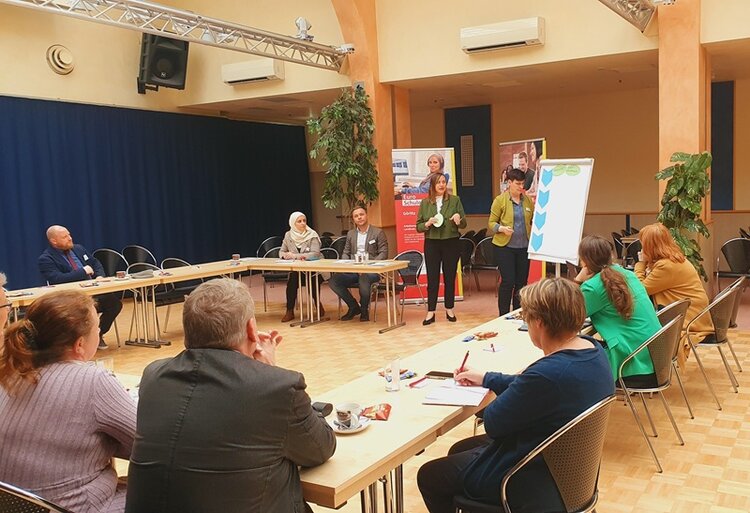 Teilnehmende sitzen an Tischen in U-Form und schauen nach vorn zu zwei Teilnehmenden, die an einem Flipchart präsentieren.