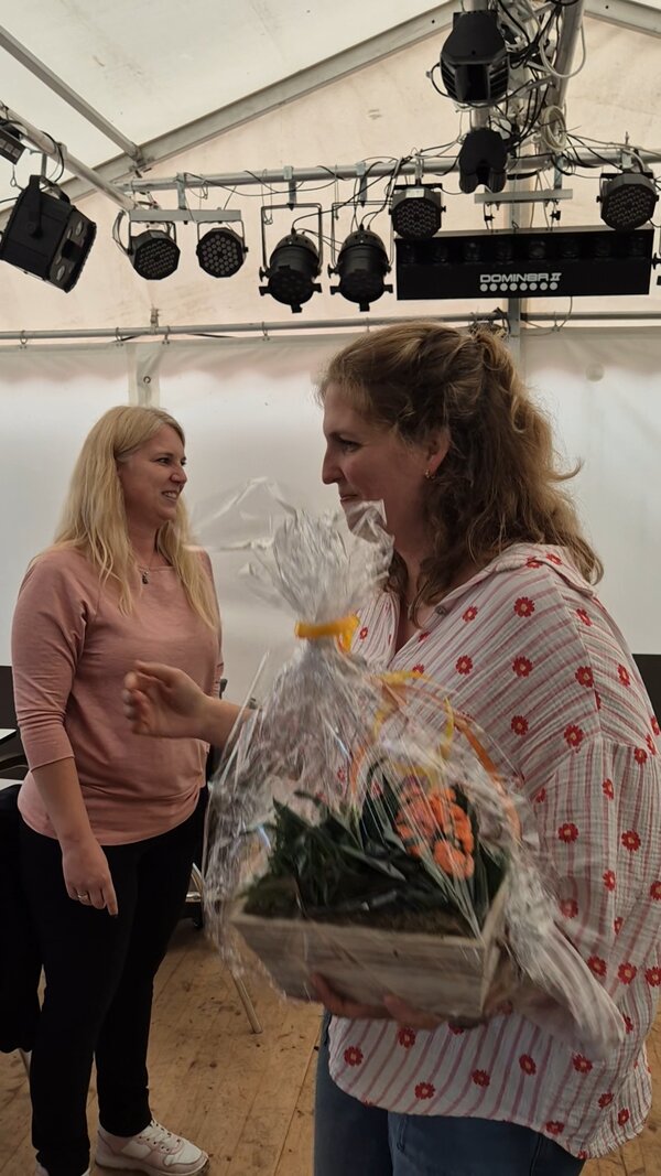 Eine scheidende Mitarbeiterin erhält zum Abschied eine Zimmerpflanze als Geschenk. Im Hintergrund steht (etwas unglücklich für die Szenerie) eine andere Mitarbeiterin, die nicht an diesem Gespräch beteiligt zu sein scheint. Das alles spielt sich in einem Festzelt ab, in dem auch eine professionelle Beleuchtungsanlage installiert ist.