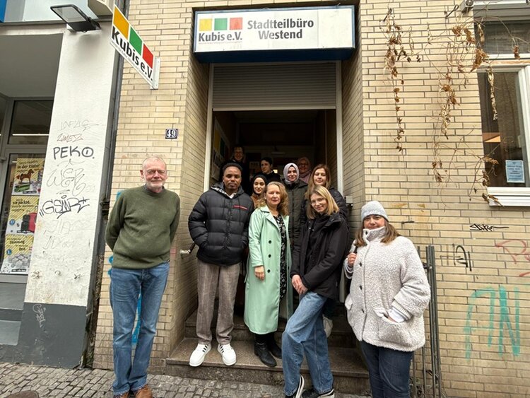 Vor einem Eingang mit dem Schild 'Kubis e.V. Stadtteilbüro Westend' stehen mehrere Teilnehmende der  Euro-Schulen Wiesbaden auf einer Treppe. Die Fassade besteht aus beigefarbenen Klinkersteinen, rechts wächst eine Kletterpflanze an der Wand. Links neben der Tür befindet sich ein weiteres Schild mit dem Schriftzug 'Kubis e.V.'. Die Personen tragen unterschiedliche Winterkleidung, einige lächeln in die Kamera. Die Hauswand ist mit Graffiti versehen.