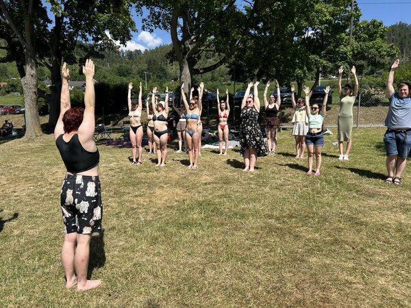 Auf einer Wiese praktiziert eine Gruppe junger Menschen Yoga: sie alle stehen und strecken die Arme gerade in die Höhe.