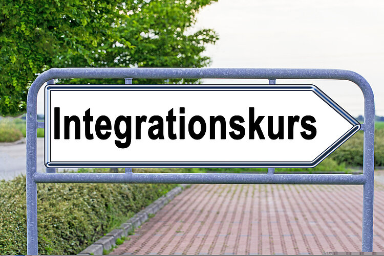 Herzlich willkommen – Neuer Integrationskurs an den Euro-Schulen Mainz startet! Ein weißes Verkehrsschild in Form eine Pfeiles, der nach rechts zeigt, weißt den Weg Richtung "Integrationskurs". Dahinter befindet sich ein gepflasterter Parkplatz.