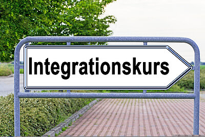 Herzlich willkommen – Neuer Integrationskurs an den Euro-Schulen Mainz startet! Ein weißes Verkehrsschild in Form eine Pfeiles, der nach rechts zeigt, weißt den Weg Richtung "Integrationskurs". Dahinter befindet sich ein gepflasterter Parkplatz.