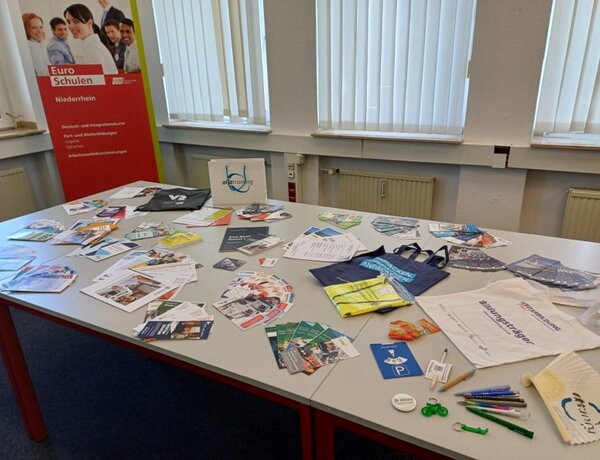 Auf zwei großen Tischen liegen verschiedene Flyer und Werbegadgets unterschiedlicher Unternehmen. In der Ecke des Raumes, hinter den Tischen steht ein rotes Roll-up der Euro-Schulen Niederrhein.