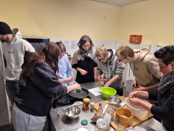 Eine Gruppe von jungen Erwachsenen arbeitet gemeinsam an einer Küchenarbeitsplatte. Sie schälen, schneiden und mischen Zutaten, während sie sich konzentriert auf ihre Aufgaben fokussieren. Verschiedene Schüsseln, Gewürze und Kochutensilien sind auf dem Tisch verteilt. Die Atmosphäre wirkt engagiert und gemeinschaftlich.