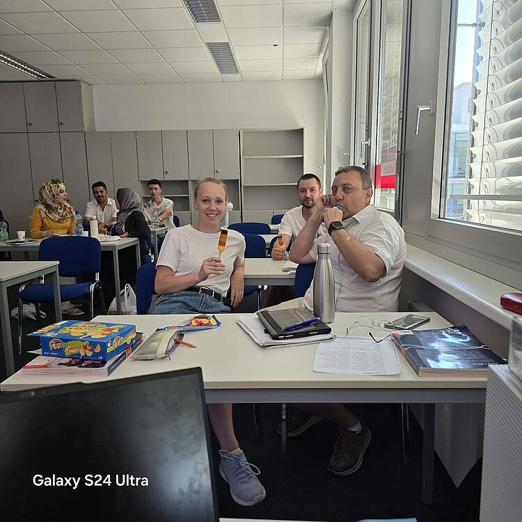 Heiße Köpfe, kühles Eis – Sprachkurse bei 35 Grad! Mehrere Kursteilnehmer sitzen in einem Klassenzimmer und essen Eis. Eine junge Frau im Vordergrund hält ein Eis am Stiel in die Kamera. Im Hintergrund unterhalten sich weitere Teilnehmer. Auf den Tischen liegen Hefte, Bücher und Getränke.