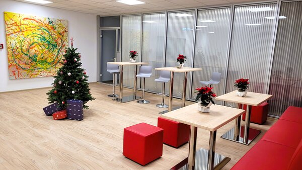 Großzügiger Aufenthaltsbereich mit hohen Stehtischen und Hockern, roten Sitzwürfeln, einem geschmückten Weihnachtsbaum mit Geschenken und mehreren Weihnachtssternen auf den Tischen; im Hintergrund eine Glaswand und ein großes buntes abstraktes Bild.