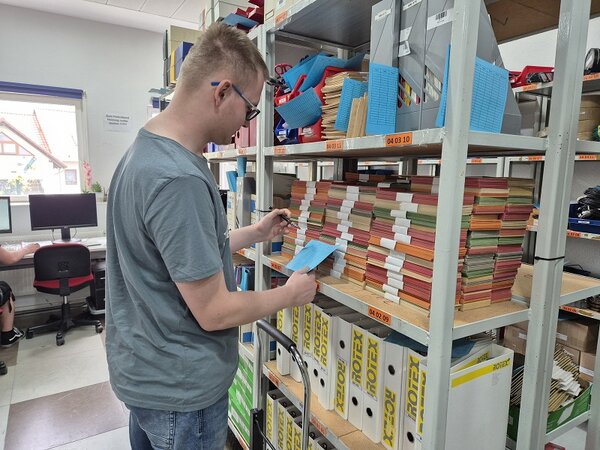 Ein Auszubildender prüft die Bestände am Lagerregal. In den Händen hält er einen Stift und eine Karteikarte.