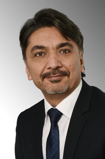 Ein Mann mit dunklem, leicht gewelltem Haar und kurzem Bart trägt einen dunklen Anzug, ein weißes Hemd und eine dunkelblaue Krawatte mit feinem Muster. Er blickt freundlich in die Kamera. Der Hintergrund ist in einem sanften Verlauf von Weiß zu Grau gehalten.