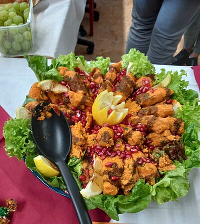 Auf einem Tisch sind auf grünen Salatblätter orientalische Köstlichkeiten drapiert. Sie sind mit Zitrone und Granatapfelkernen dekoriert. Ein Löffel zum Rausnehmen liegt parat. Hinten im Bild kann man noch eine Schüssel mit Trauben erkennen.