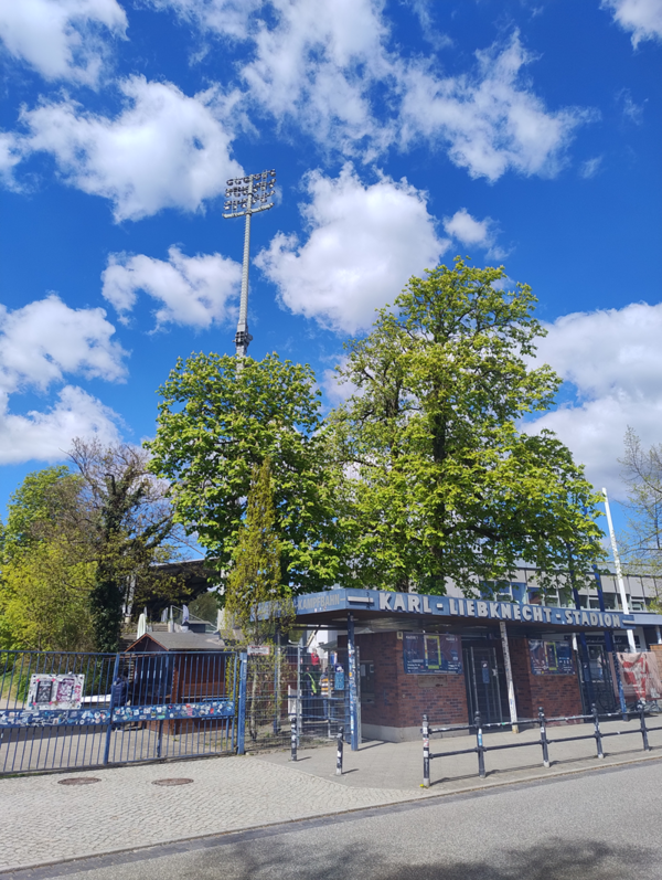 Vor dem Eingang eines Stadions mit dem Schriftzug 'KARL-LIEBKNECHT-STADION' stehen mehrere hohe Bäume mit frischem Laub. Im Hintergrund ragt ein Flutlichtmast in den blauen Himmel mit weißen Wolken. Der Eingangsbereich besteht aus einem Backsteingebäude mit Metalltoren und einem Zaun. Auf dem Gehweg davor sind Poller aufgestellt.