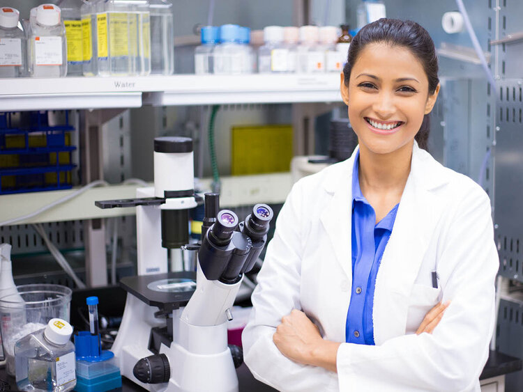 Eine lächelnde Wissenschaftlerin in einem weißen Laborkittel steht mit verschränkten Armen in einem Labor. Im Hintergrund sind Laborgeräte wie ein Mikroskop, Reagenzgläser und Chemikalienflaschen zu sehen.