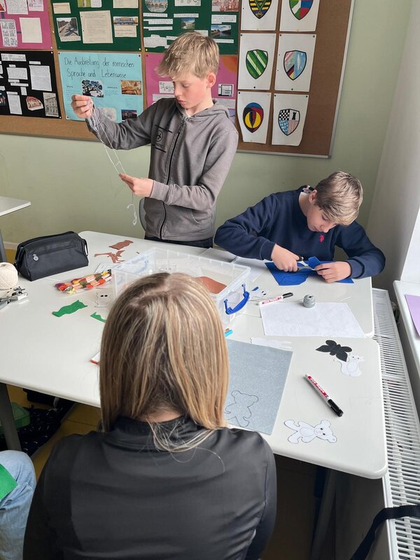 Drei Schüler*innen arbeiten an einem Bastelprojekt. Ein Junge entwirrt Wolle, ein anderer schneidet aus Filz, während auf dem Tisch Tierfiguren und Bastelmaterialien liegen.