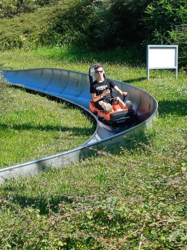 Ein junger Mann mit reflektierender Sonnenbrille bremst mit einer Hand das Gefährt einer Sommerrodelbahn. In der anderen Hand hält er ein Handy.