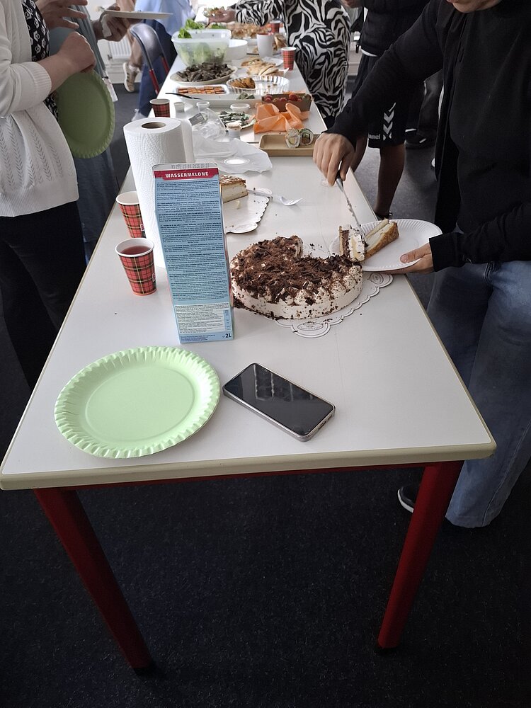 Sternstunde im Leben eines Deutschlehrers Ein Buffet-Tisch mit verschiedenen Speisen; im Vordergrund wird eine Torte angeschnitten, während mehrere Personen Teller und Essen nehmen.