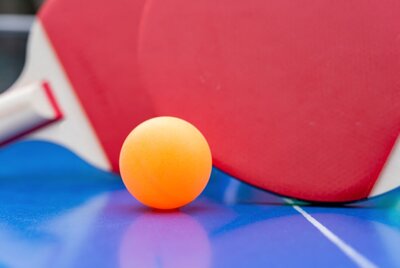 Ein orangefarbener Tischtennisball liegt auf einem blauen Tischtennistisch, im Hintergrund sind zwei rote Tischtennisschläger zu sehen.