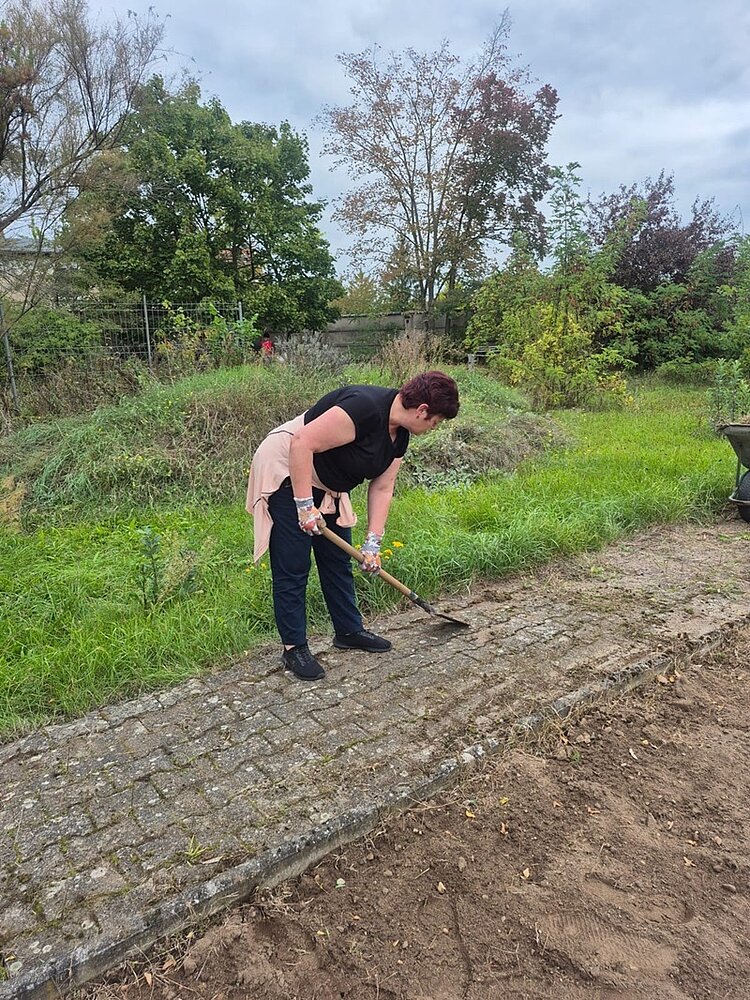 Eine Teilnehmende entfernt mithilfe eines Spatens Unkraut in den Rillen des Gehweges. Sie trägt eine schwarze Hose, ein schwarzes T-Shirt, einen um die Hüfte gewickelten hellen Pullover und eine dunkle Kurzhaarfrisur.