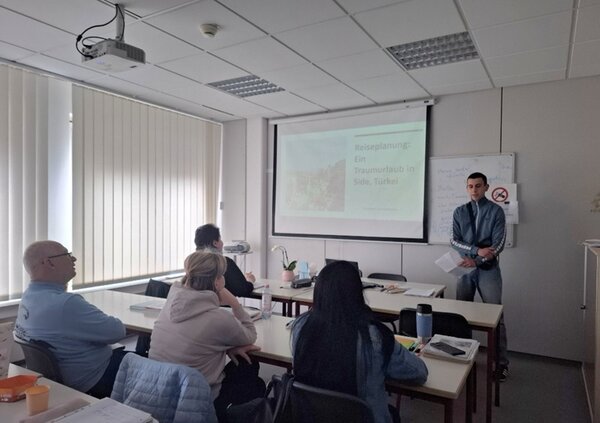 Ein junger männlicher Teilnehmer in Jeanshose und Sportjacke steht vorn am Whiteboard im Unterrichtsraum und hält seine Präsentation vor seinen Mitteilnehmenden des B2-Sprachkurses an den Euro-Schulen Magdeburg.