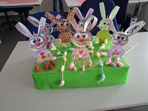 Auf einem grünen Karton sitzen mehrere aus buntem Papier gebastelte Osterhasen.