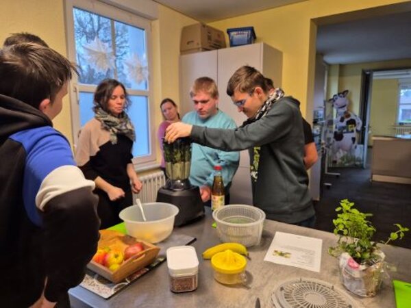 Eine Gruppe von Jugendlichen und Erwachsenen steht in einer Küche um eine Arbeitsplatte. Ein junger Mann bedient einen Mixer, in den frische grüne Zutaten gegeben werden. Auf dem Tisch liegen verschiedene Lebensmittel wie Äpfel, Zitronen und Kräuter sowie ein Rezeptblatt. Im Hintergrund sind weitere Teilnehmerinnen und Teilnehmer sowie eine dekorierte Wand mit einer Kuhfigur aus Pappe zu sehen. Die Atmosphäre wirkt konzentriert und gemeinschaftlich.