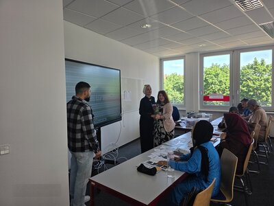 Die Teilnehmenden des Integrationskurses an den Euro-Schulen Dresden sitzen an Tischen, einige stehen vorn am Whiteboard mit der ehemaligen Teilnehmerin und überreichen ihr einen Blumenstrauß.