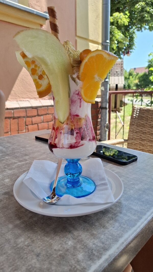 Großer Eisbecher mit Sahne, Fruchtsoße, Melone, Orange, Physalis und einer Waffelröllchen-Garnitur auf einem Außentisch.