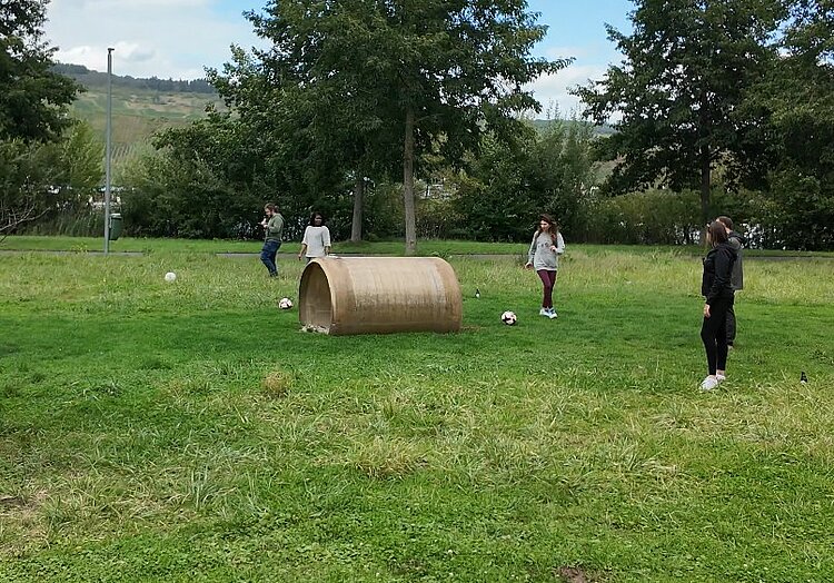 Team-Building beim Fußballgolf In der Mitte der Wiese liegt ein Rohr. Die Mitarbeitenden der Euro-Schulen Trier stehen mit ihren Bälle außenherum und machen offenbar einen Schlachtplan für das weitere Vorgehen.