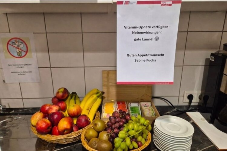 Auf einer Küchenarbeitsplatte stehen zwei Obstkörbe mit Äpfeln, Bananen, Kiwis sowie grünen und roten Trauben; daneben eine Teebeutel-Auswahl und gestapelte Teller. Darüber hängt ein Aushang: „Vitamin-Update verfügbar – Nebenwirkungen: gute Laune! Guten Appetit wünscht Sabine Fuchs“.